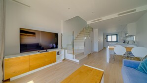 Un salon moderne doté d’un téléviseur à écran plat, d’un meuble en bois et d’un escalier menant à un espace cuisine comprenant une table et des chaises.