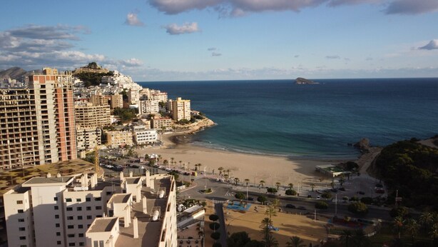 A coastal cityscape with high-rise buildings, a beach, and a rocky outcrop in the distance.