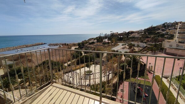 A balcony with a view of a coastal town, a sandy area with palm trees, and a clear blue sky.