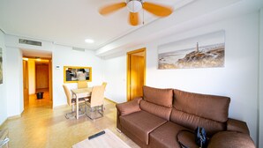 A living room with a brown sofa, a dining table with chairs, a ceiling fan, and a painting of a lighthouse on the wall. - AL Piso de 2 dormitorios en El Campello (El Campello)
