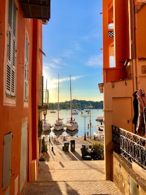 Una calle estrecha que lleva a un puerto con barcos, con un edificio rojo con contraventanas y un balcón.