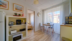 Una cocina compacta con comedor, que incluye microondas, hervidor de agua y cafetera en estantes, una mesa pequeña con sillas y un ventanal con cortinas.