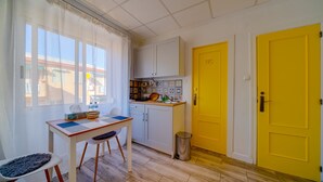 Una cocina compacta con armarios blancos, una puerta amarilla con la inscripción 'WC', un pequeño cubo de basura y una mesa con sillas.