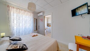 Habitación de hotel con cama, escritorio, televisión y una cocina visible a través de una puerta abierta.