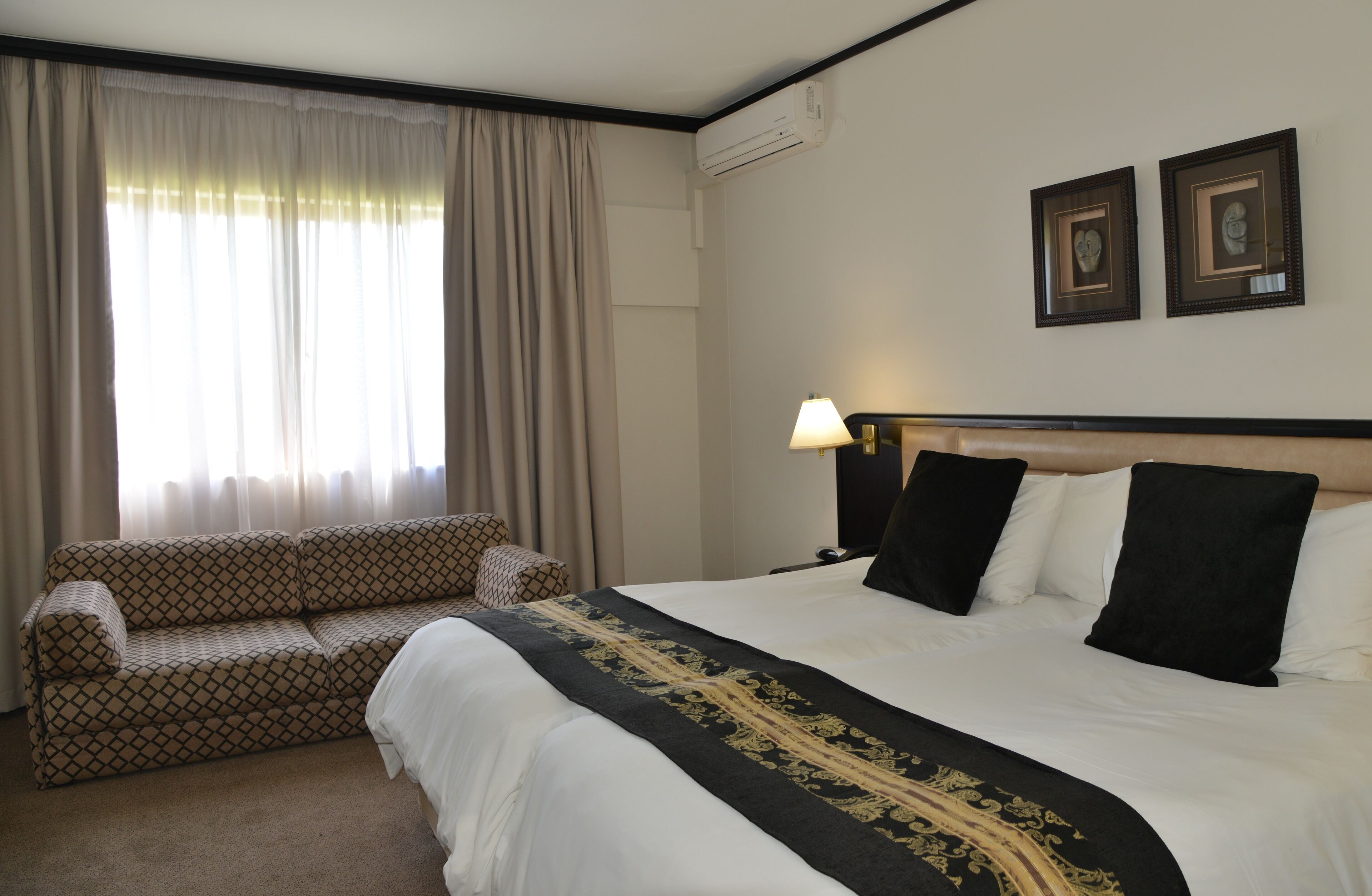 Quarto de hotel com uma cama, um sofá, uma luminária e duas fotos emolduradas na parede.