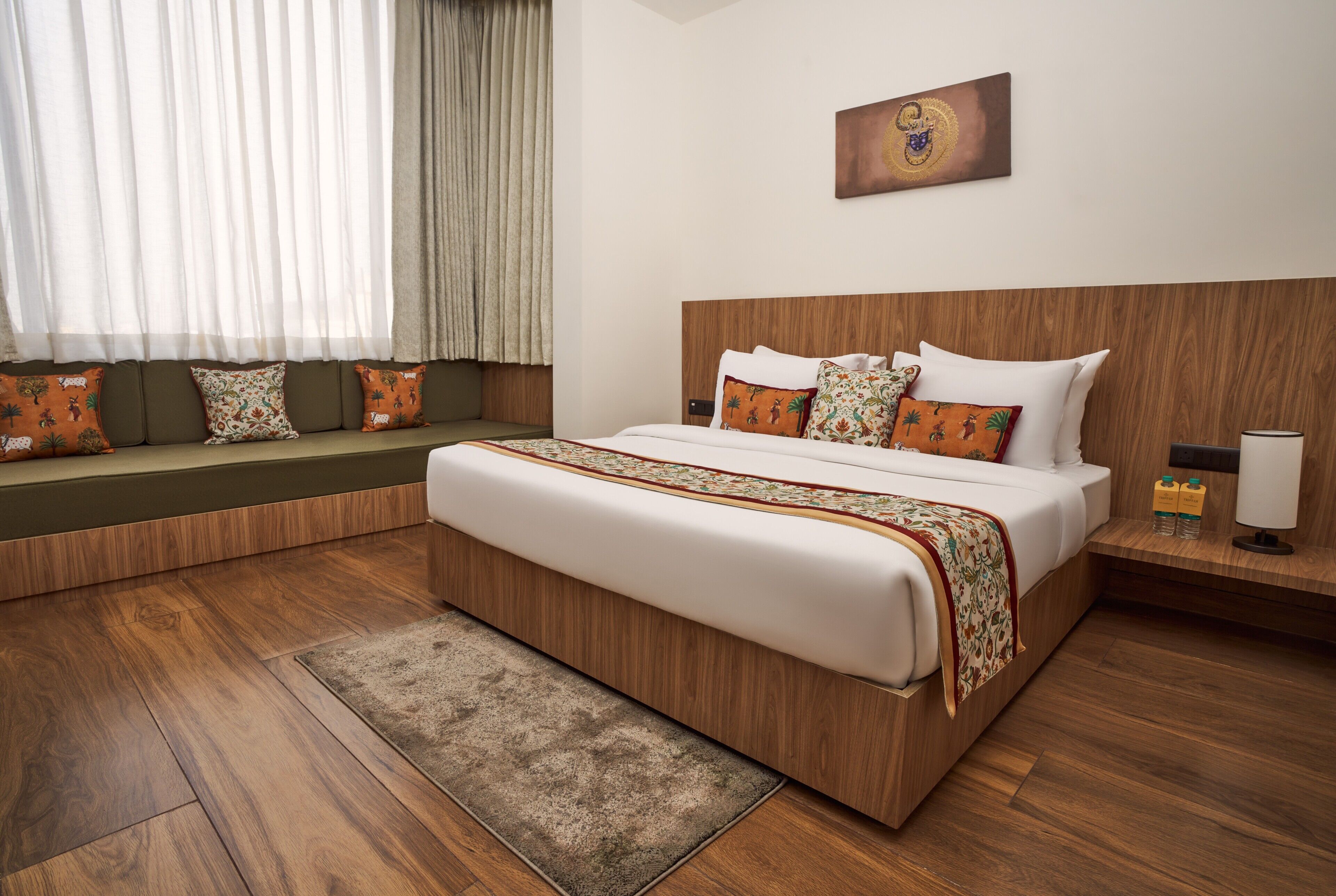 A hotel room with a large bed, wooden headboard, a bench with cushions, a wooden floor, and a window with curtains.