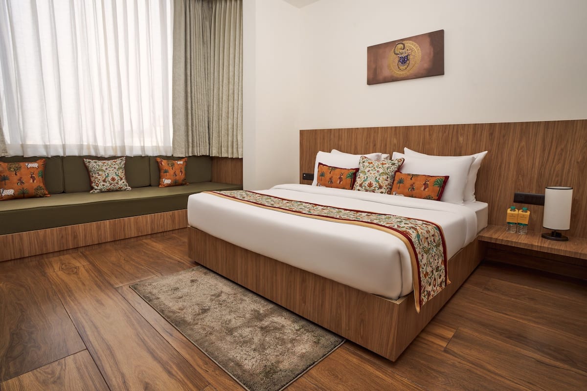 A hotel room with a large bed, wooden headboard, a bench with cushions, a wooden floor, and a window with curtains.
