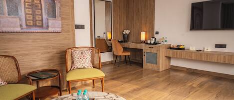 A hotel room with a wooden floor, a marble table, wicker chairs, a desk with a flat-screen TV, and a decorative wall with a mirror.