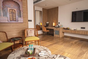 A hotel room with a wooden floor, a marble table, wicker chairs, a desk with a flat-screen TV, and a decorative wall with a mirror.