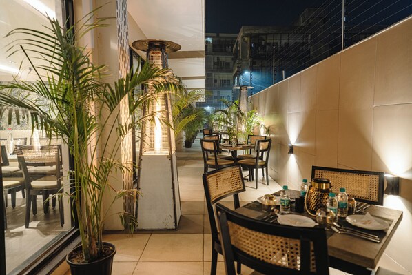 An outdoor dining area with tables and chairs, potted plants, and a tall, illuminated column.