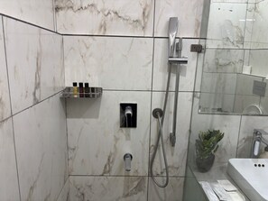 A bathroom with a shower, a glass shelf with toiletries, a potted plant, and a sink.
