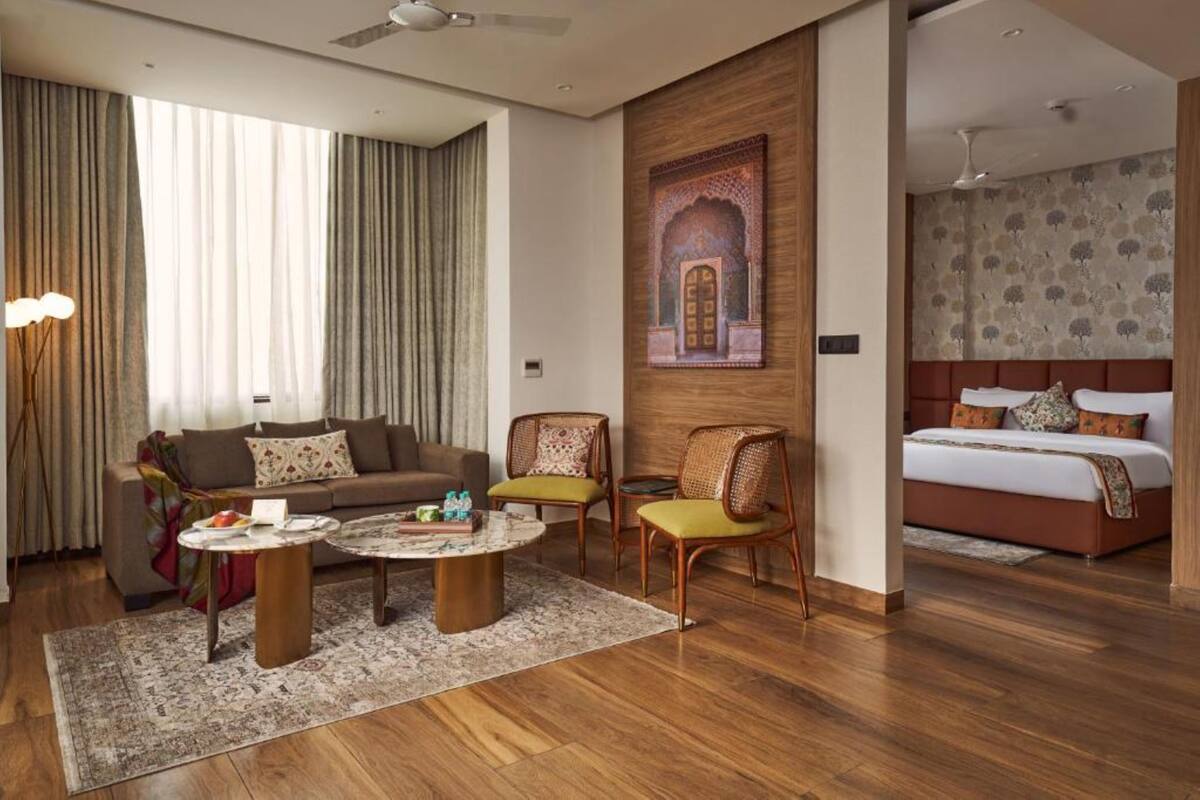 A modern hotel room with a sofa, two armchairs, a coffee table, a bed, and a large window with curtains.