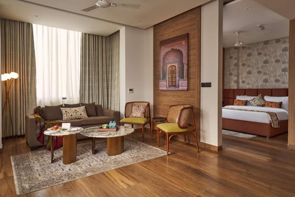 A modern hotel room with a sofa, two armchairs, a coffee table, a bed, and a large window with curtains.