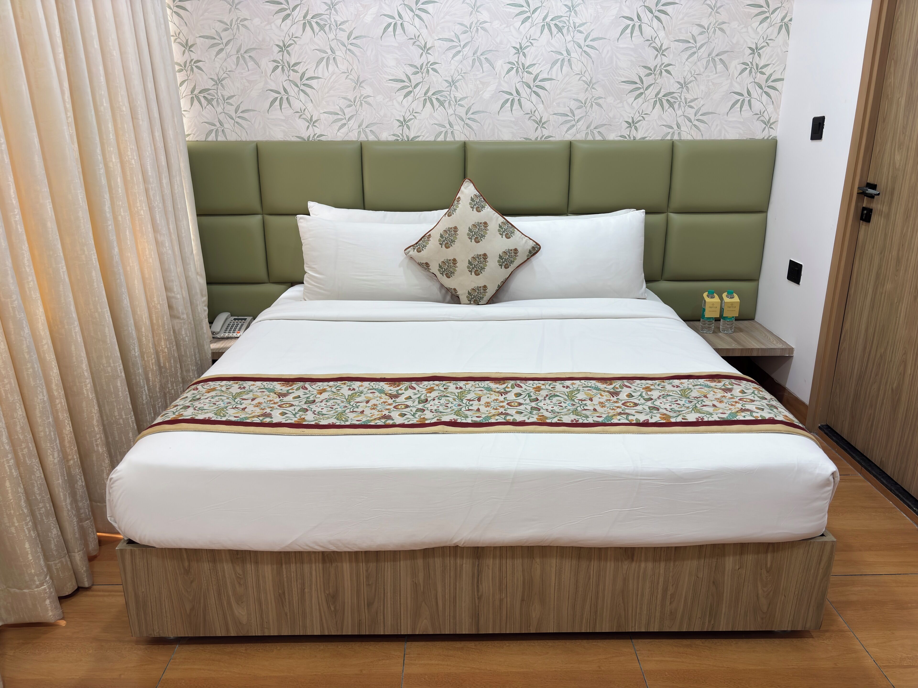 A hotel room with a green upholstered headboard, white bedding, a patterned throw, and a bedside table with water bottles.