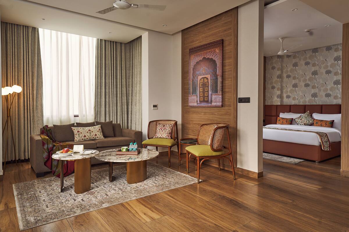 A modern hotel room with a sofa, two armchairs, a round coffee table, a bed with a headboard, and a large window with curtains.