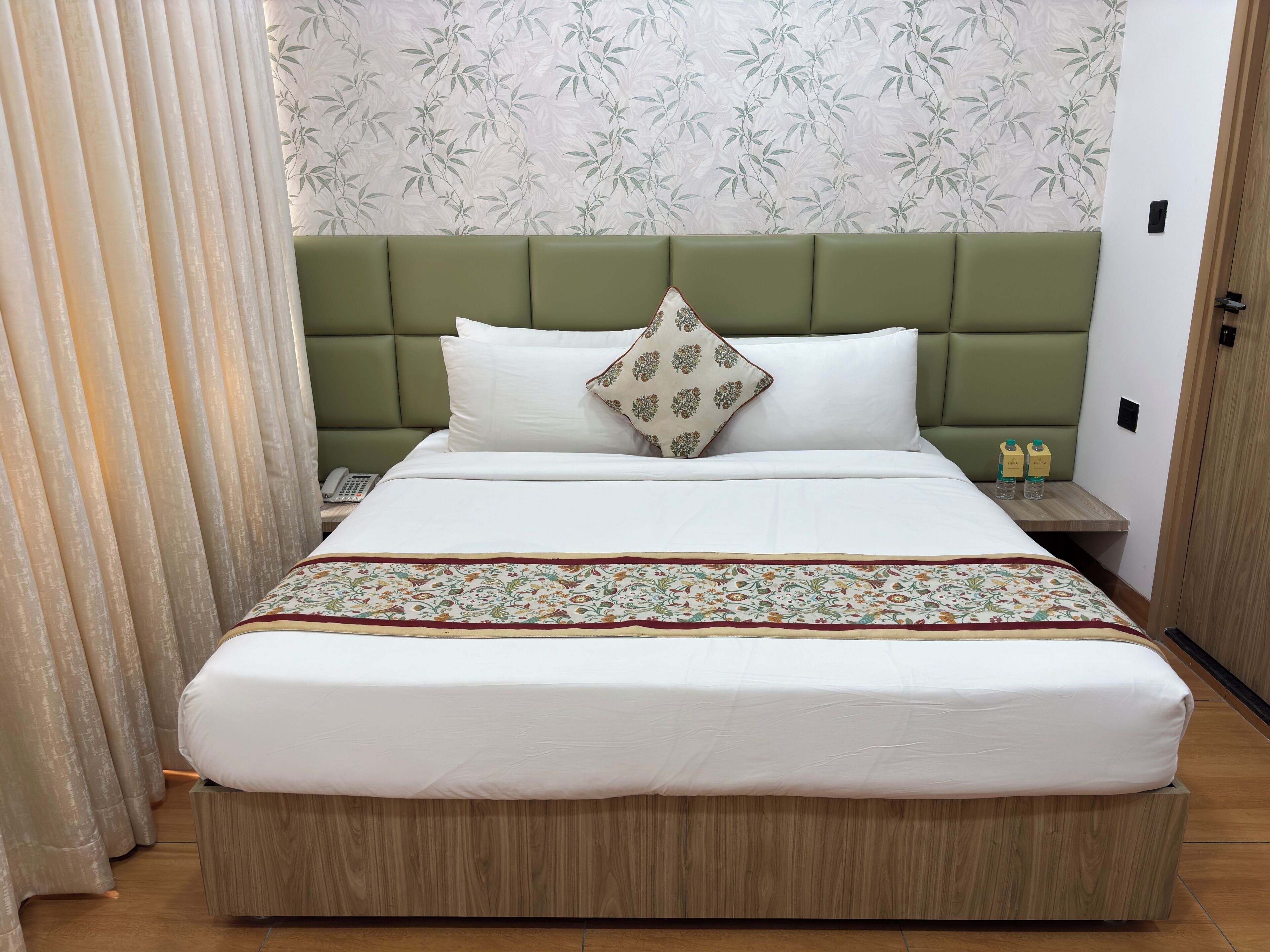 A neatly made bed with a green headboard, a patterned bedspread, and two bottles on a nightstand.