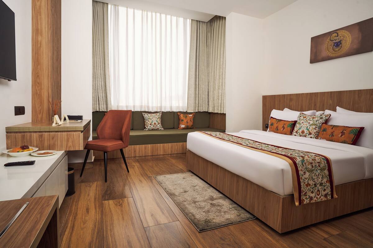 A hotel room with a bed, a desk, a chair, a window with curtains, and a wall-mounted TV.