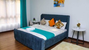 A bedroom with a bed, a small table, a chair, and a painting on the wall. - Lux suites Rosewood Park Apartments (Nairobi)