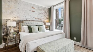 Une chambre avec un grand lit, un banc, une petite table et un mur en briques.
