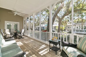 A covered porch with a ceiling fan, a dining table with chairs, a sofa, and a small table with a vase and lemons.