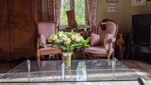 A room with a glass-top table, two striped armchairs, a wooden cabinet, and a vase with white flowers.