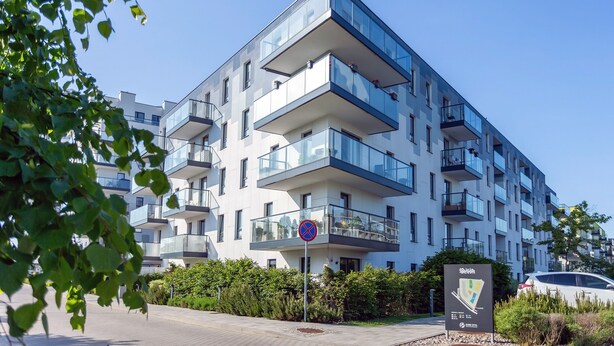 A modern multi-story residential building with balconies, a clear sky, and a parking area with a car.