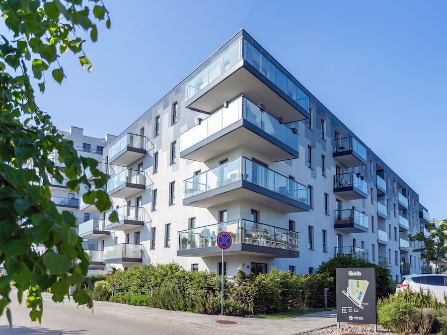A modern multi-story residential building with balconies, a clear sky, and a parking area with a car.