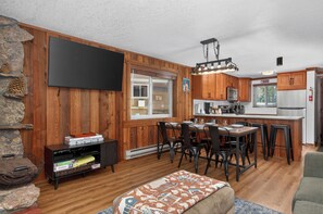 A cozy living room with a fireplace, wooden walls, a flat-screen TV, and a dining area with a kitchen in the background. - Family friendly 2 bedroom cabin themed interior (Winter Park)