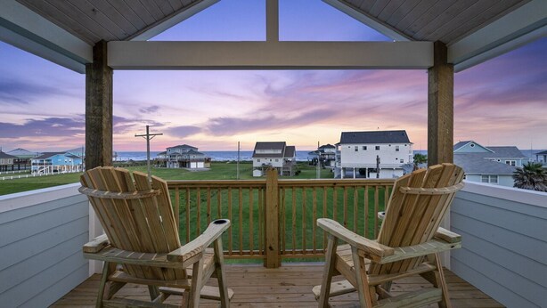 A wooden deck with two Adirondack chairs facing a scenic view of houses and the ocean at sunset.