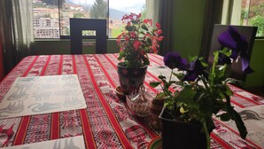 Een kamer met een eettafel, een raam met uitzicht op gebouwen en bomen, en een potplant met rode bloemen.