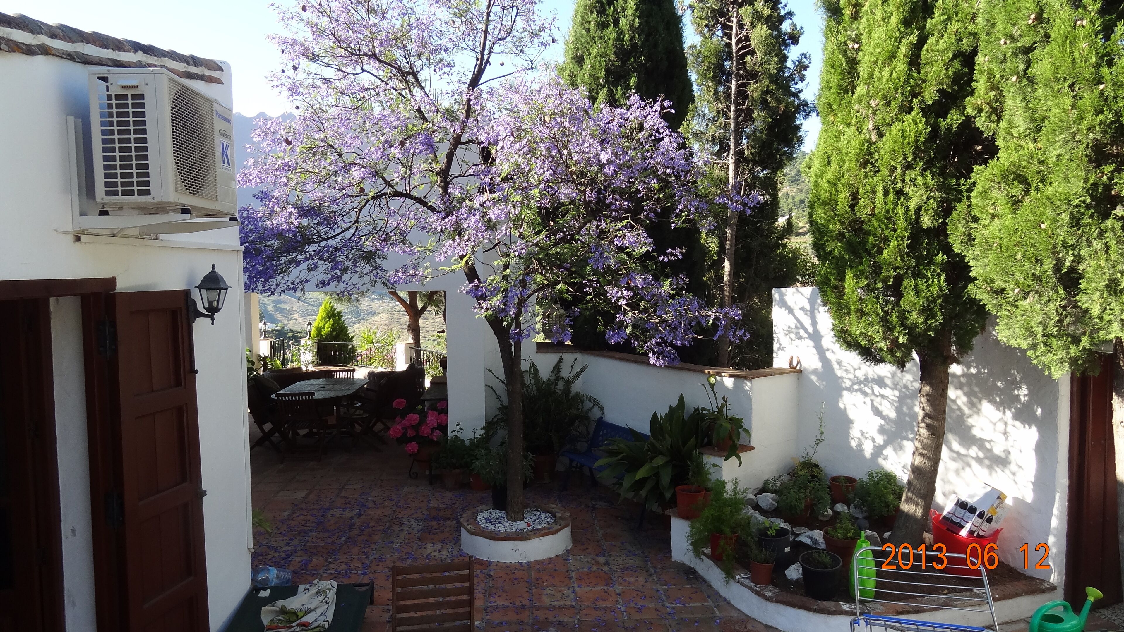 Eine Terrasse im Freien mit Tisch und Stühlen, umgeben von Topfpflanzen und einem blühenden Baum.