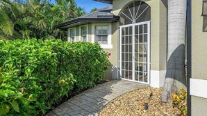 A house with a large window, a covered porch, and a well-maintained garden with a hedge and palm trees. - Kings Castle - 3BR w/ Bikes & Boat Dock Access (Cape Coral)