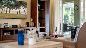 A living room with a large TV, a wooden coffee table, a glass of wine, and a bottle of wine. There is a wooden cabinet with a picture frame and a clock on the wall. - Kings Castle - 3BR w/ Bikes & Boat Dock Access (Cape Coral)
