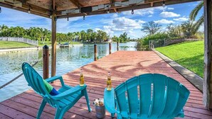 A wooden deck with two turquoise chairs and a table, overlooking a calm body of water with a dock and greenery in the background. - Kings Castle - 3BR w/ Bikes & Boat Dock Access (Cape Coral)