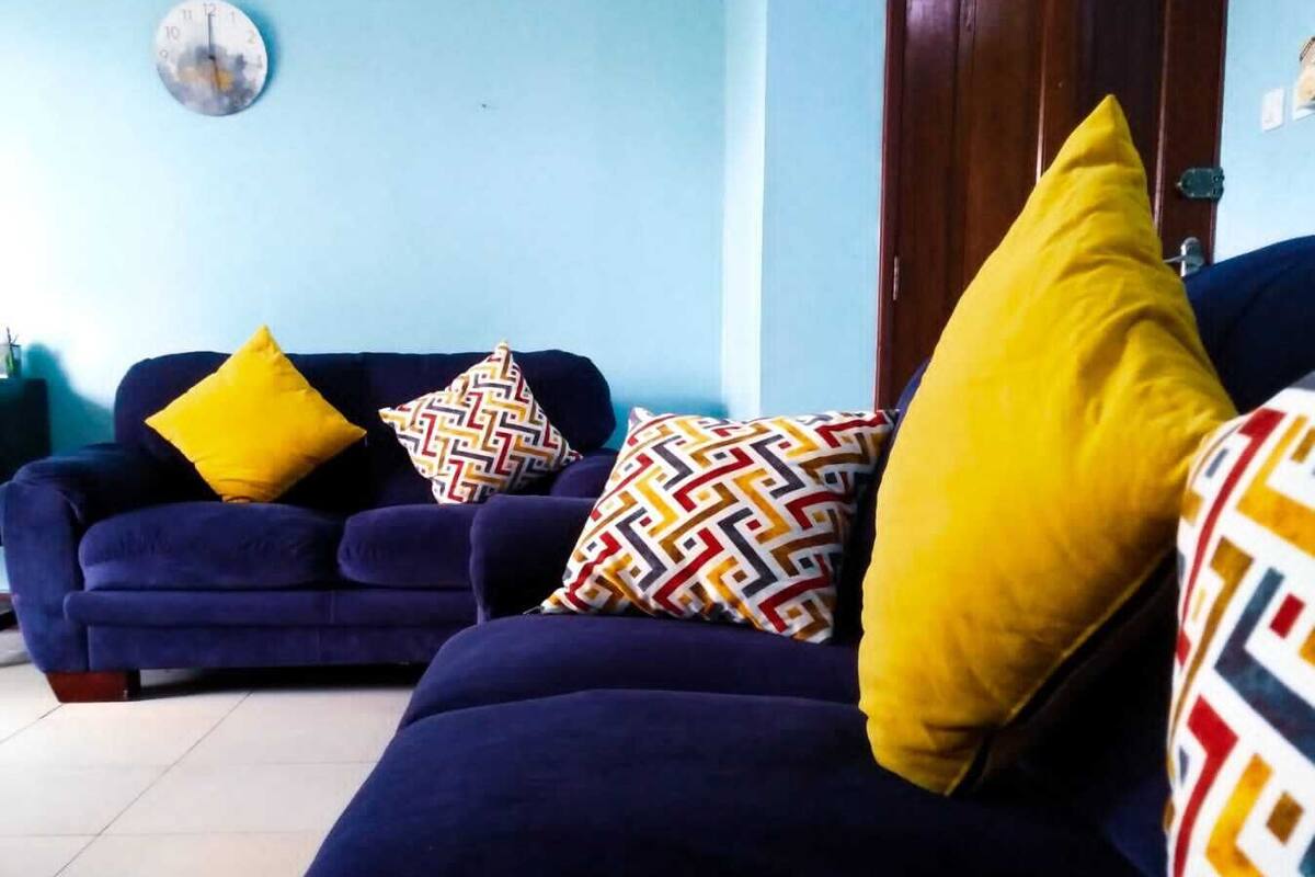 A living room with a dark blue sofa set, yellow and patterned cushions, a clock on the wall, and a wooden door.
