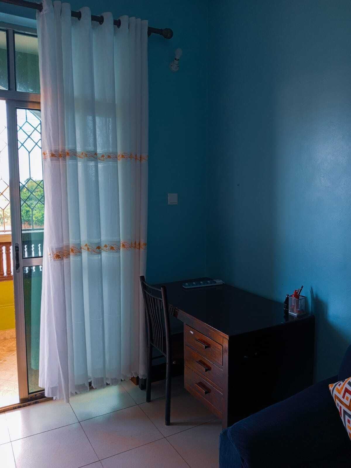 A room with a desk, chair, and a view through a window with a curtain.