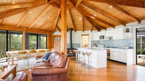 A spacious living area with a wooden ceiling, a leather sofa, a dining table with chairs, and a kitchen with white cabinets and a blue backsplash.