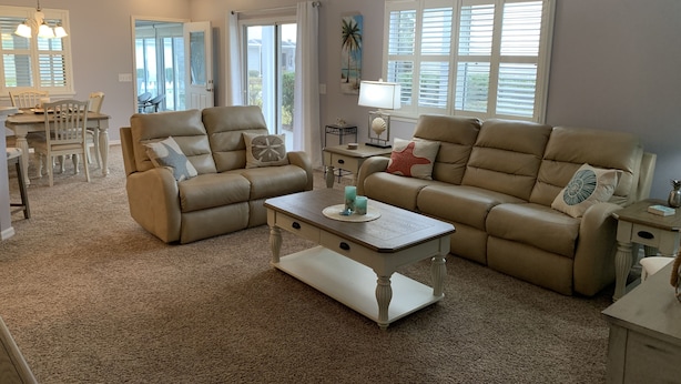 A spacious living room with a beige sofa, a matching armchair, and a coffee table. There is a dining area with a table and chairs, and a window with blinds. - Central Location!! Beautiful Villa with 2 Seater Gas Golf Cart & WIFI (The Villages)