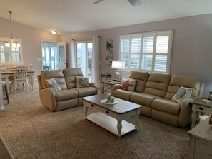 Ein geräumiges Wohnzimmer mit einem beigefarbenen Sofa, einem passenden Sessel und einem Couchtisch. Es gibt eine Essecke mit Tisch und Stühlen sowie ein Fenster mit Jalousien.