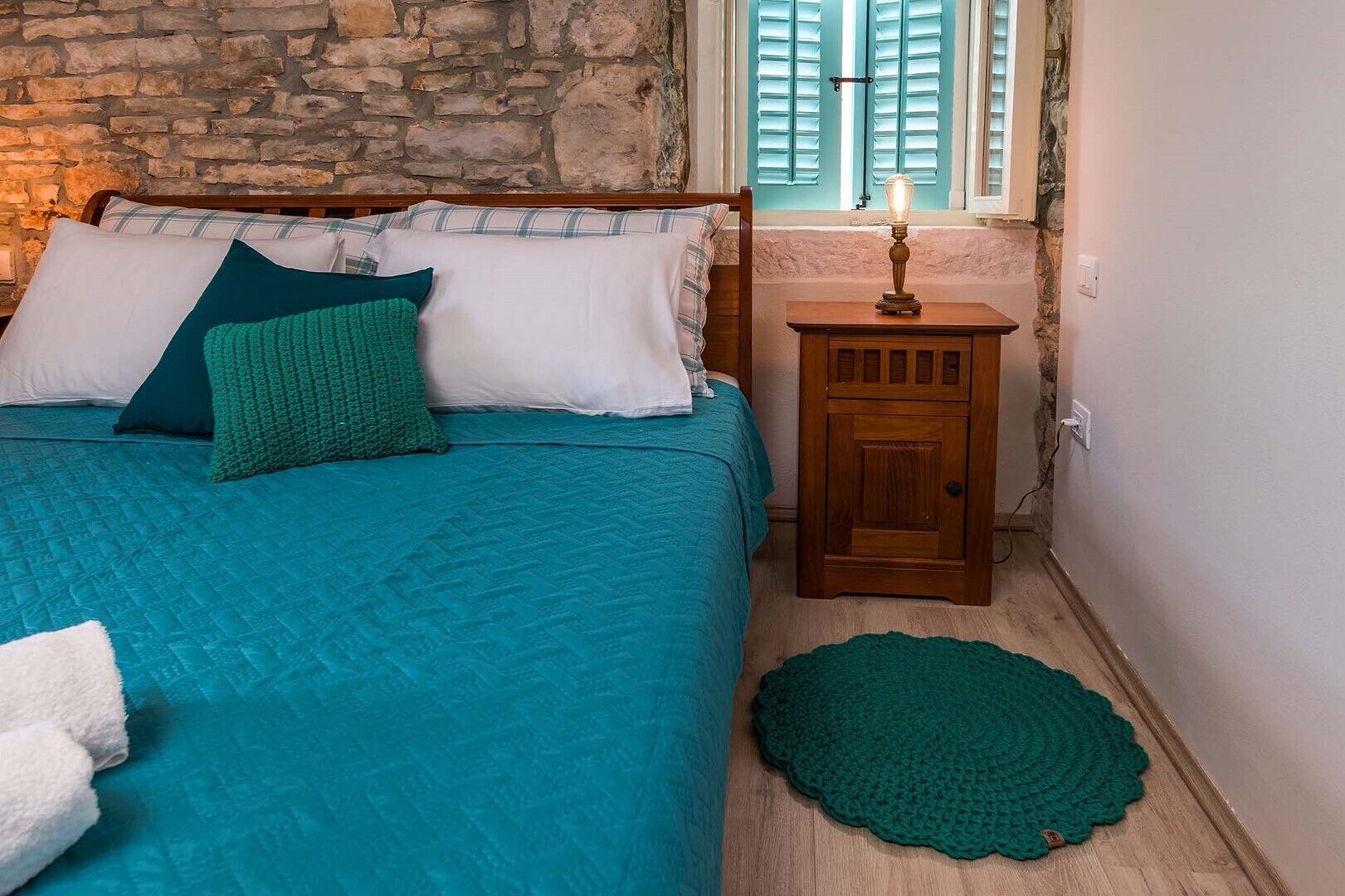 Una camera da letto con una parete in pietra, un letto con biancheria color petrolio, un comodino con una lampada e un tappeto verde all'uncinetto.
