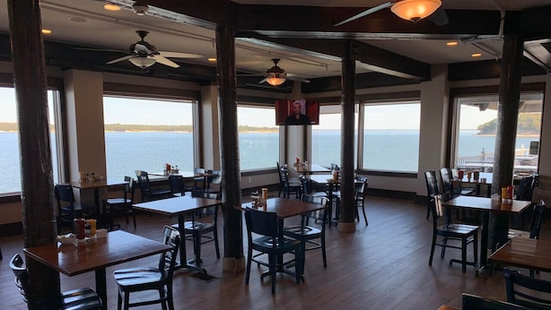 Un restaurante con mesas y sillas de madera, amplias ventanas con vista al mar y ventiladores de techo.
