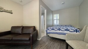 Ein Schlafzimmer mit einem braunen Ledersofa, einem beige Armlehnstuhl, einem Bett mit blauen, gemusterten Bettwäsche und einem Fenster mit Jalousien.