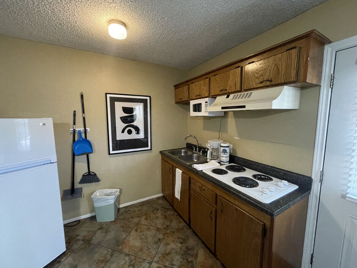 Une cuisine de taille réduite, avec un réfrigérateur blanc, une cuisinière, un four à micro-ondes, des placards en bois et un balai.