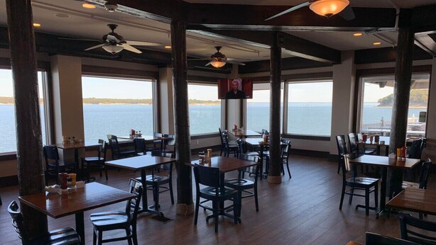 Un restaurant avec des tables et des chaises en bois, de grandes fenêtres donnant sur la mer et des ventilateurs de plafond.
