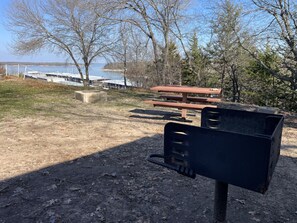 Ein Picknickplatz mit Bank und Grill, umgeben von Bäumen und einem Gewässer.