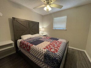 Ein Schlafzimmer mit Bett, Nachttisch, Wandregal, Deckenventilator und Fenster mit Jalousien.