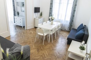Un salon moderne avec un parquet en bois à motifs chevrons, une table à manger blanche avec des chaises, un canapé foncé agrémenté de coussins bleus et une petite table d’appoint blanche sur laquelle est posée une plante.