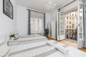 Ett hotellrum med två sängar, en kristallkrona, en balkong med utsikt över byggnader och ett fönster med gardiner.