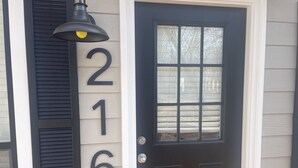 Une porte d’entrée noire avec un panneau de verre, flanquée d’un luminaire et d’une plaque indiquant le numéro de la maison.