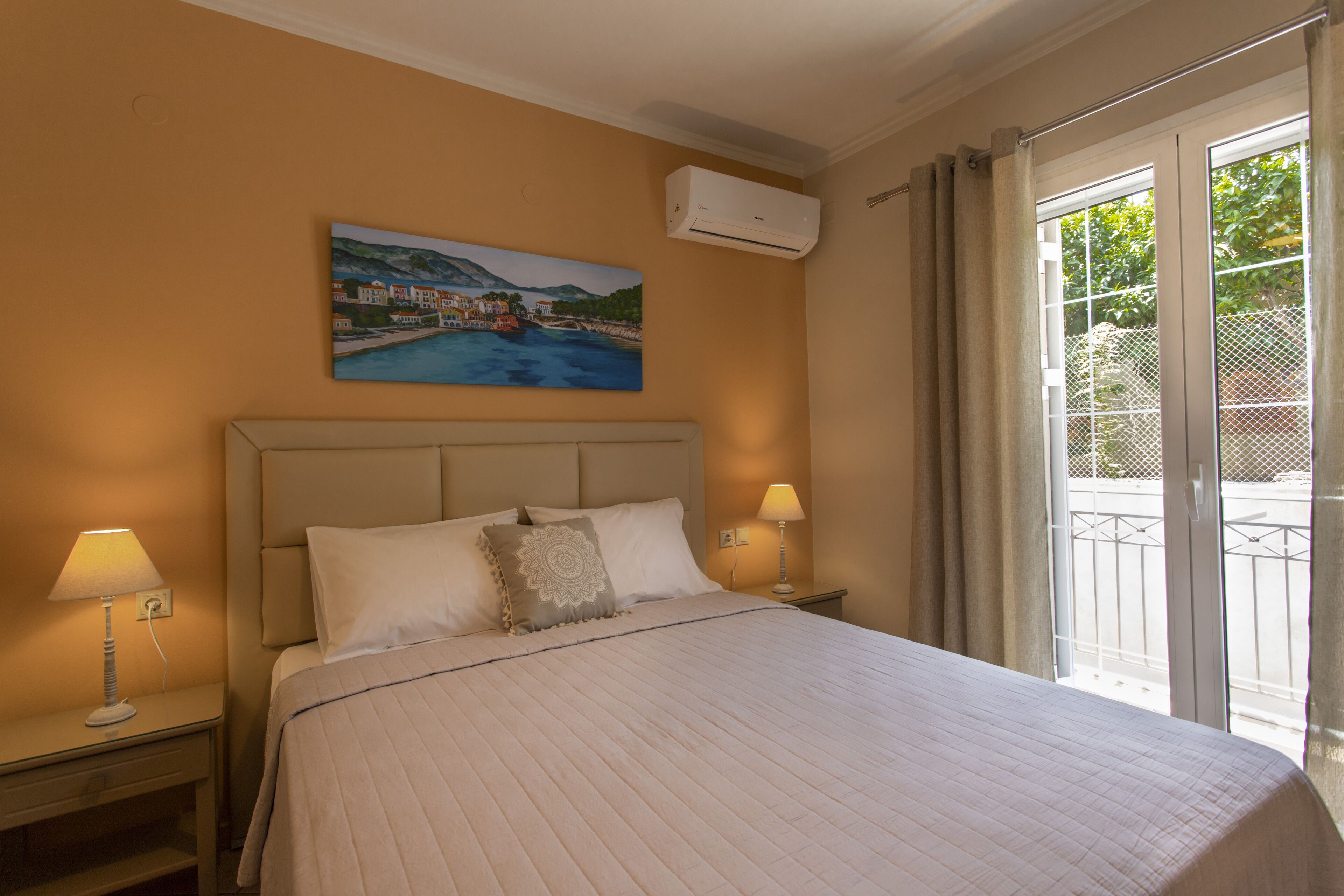 Une chambre d’hôtel avec un grand lit, deux lampes de chevet, un tableau représentant un paysage côtier, un appareil de climatisation et une porte-fenêtre coulissante donnant sur l’extérieur.